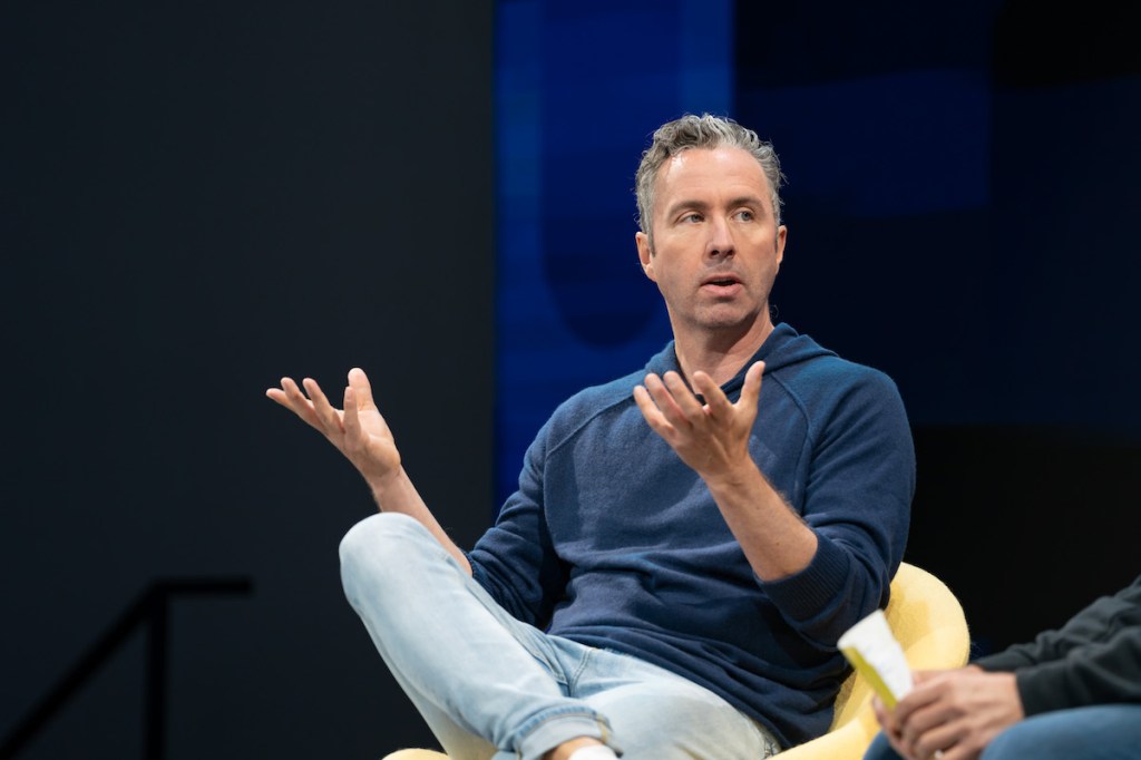 A man sitting in a yellow chair on stage and gesturing with his hands.