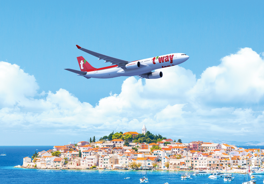 A T'way aircraft flying over a European seaside town.