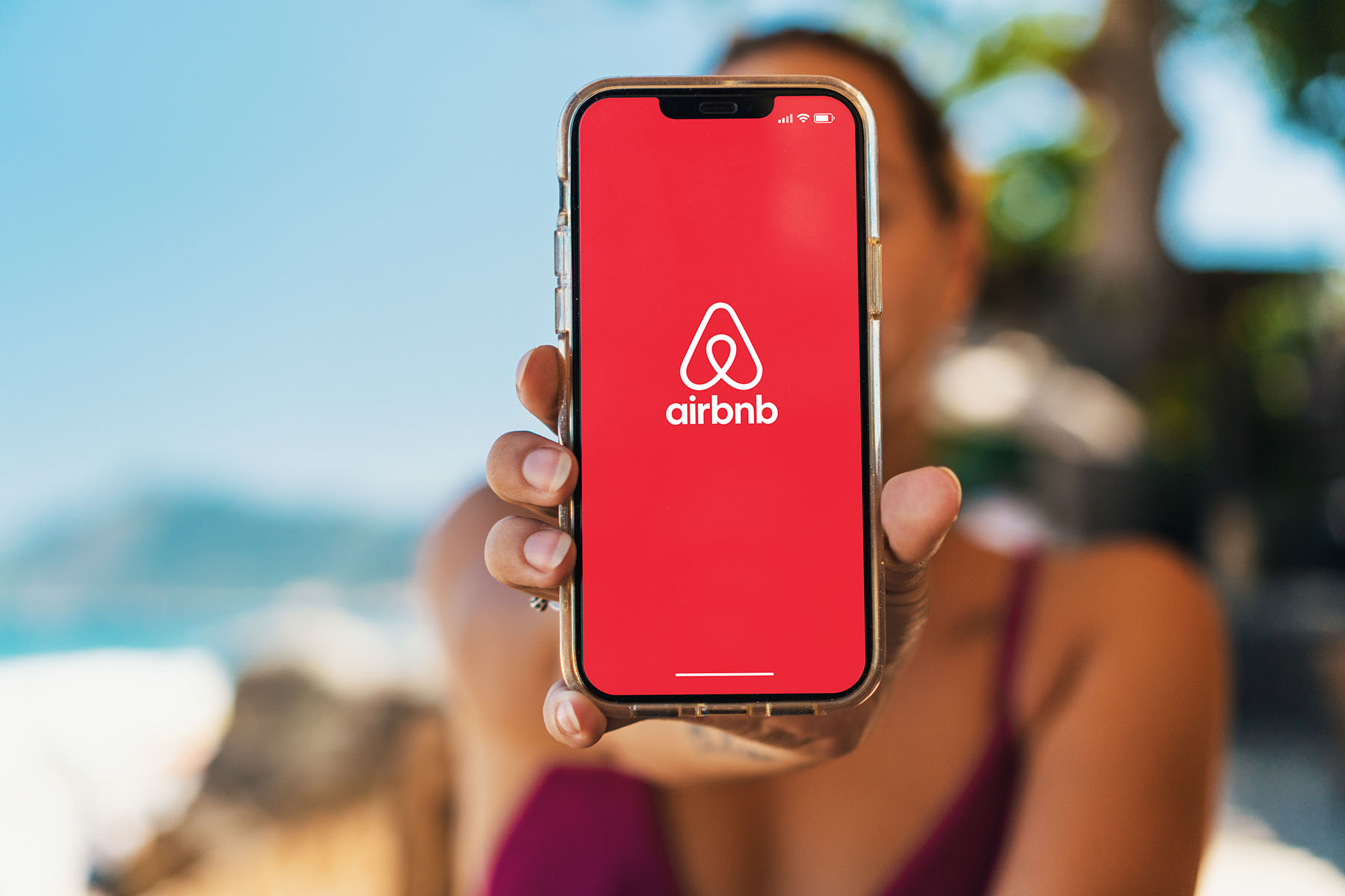 Woman on a beach holding a smartphone with Airbnb app on the screen.