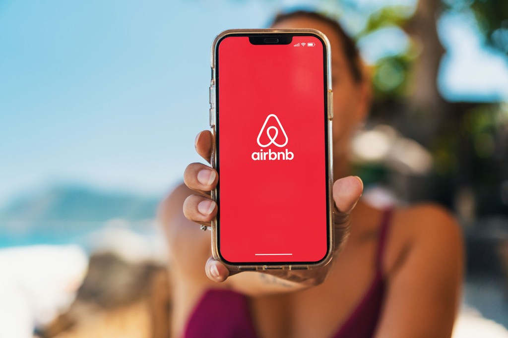 Woman on a beach holding a smartphone with Airbnb app on the screen.
