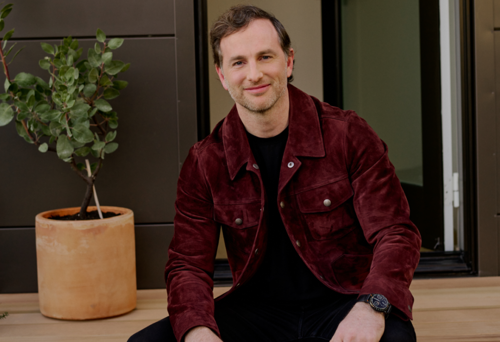 A man seated with a plant in the background.