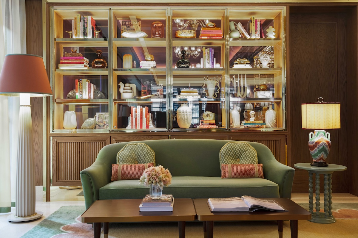 A hotel lobby with a colorful shelf filled with books and artistic pieces. A green couch and brown table are in the foreground.