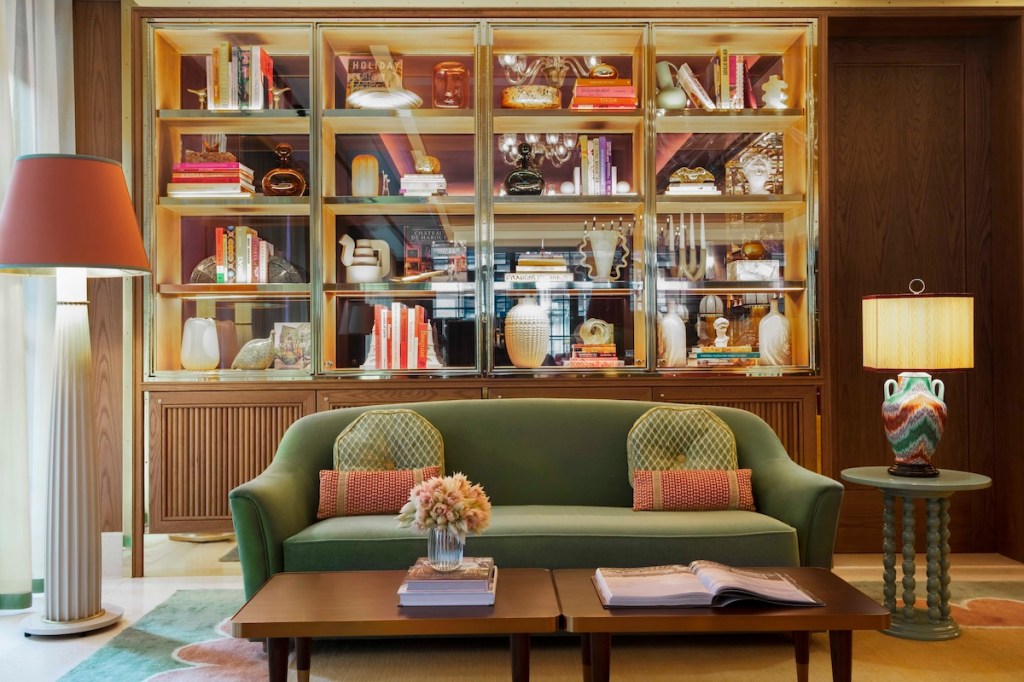 A hotel lobby with a colorful shelf filled with books and artistic pieces. A green couch and brown table are in the foreground.