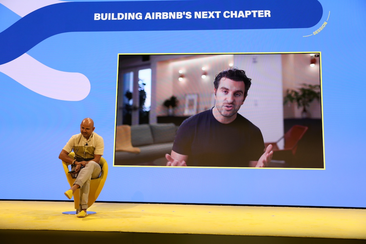 Airbnb CEO Brian Chesky in the background appearing virtually. Skift founder Rafat Ali in the foreground sitting on a yellow chair.