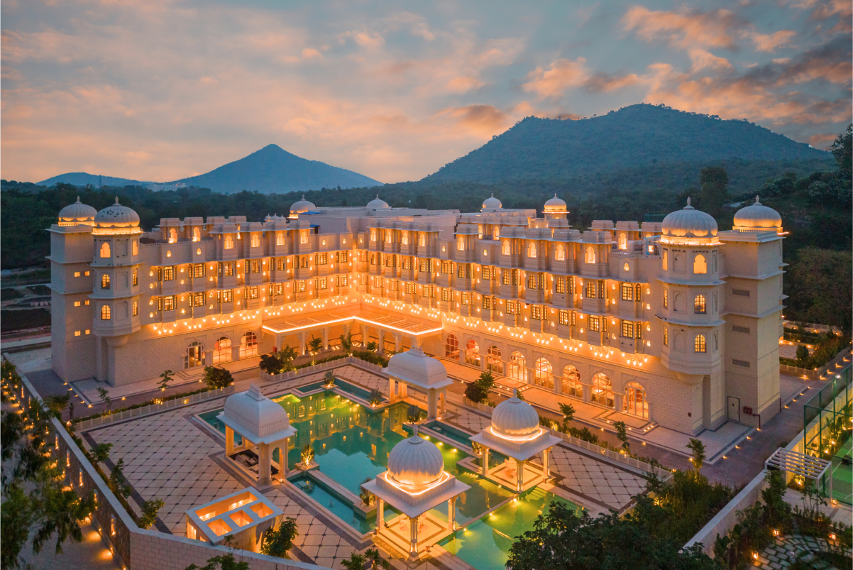 Nestled against the majestic Aravalli mountains a brightly lit Indian hotel is shown with its pool at sunset