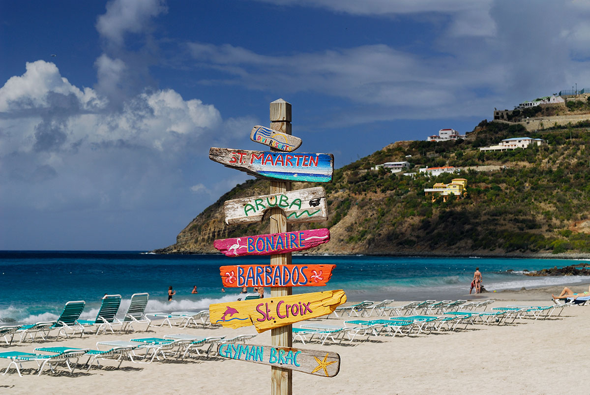 Colorful signpost of Caribbean island names on a beach.