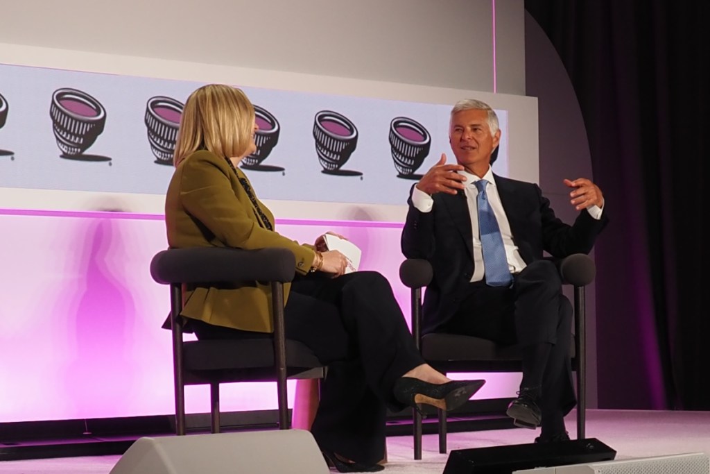 view of a ceo in a dark business suit talking on a stage at a conference with a stage moderator