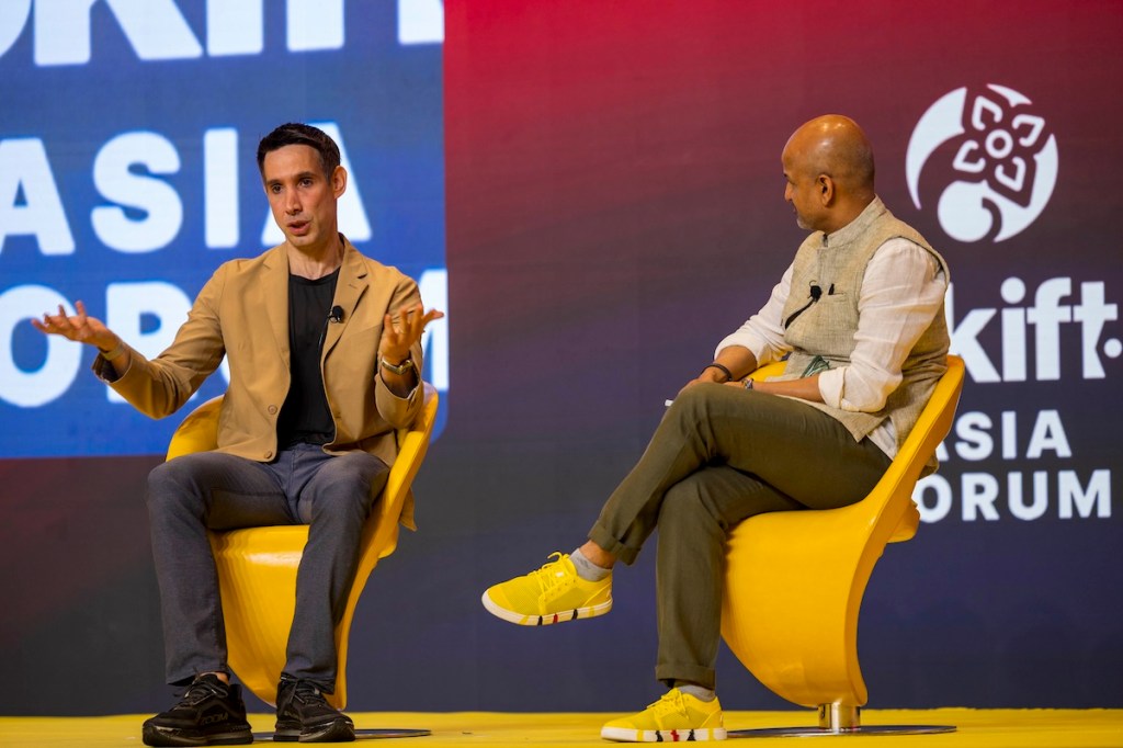 two men sitting on stage, one is a sport coat and one wearing a vest.