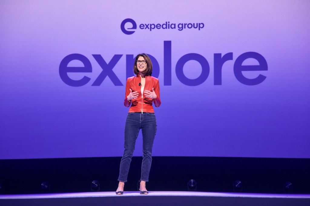 A woman in a red leather jacket standing on a stage and the words Expedia Explore are in the background.