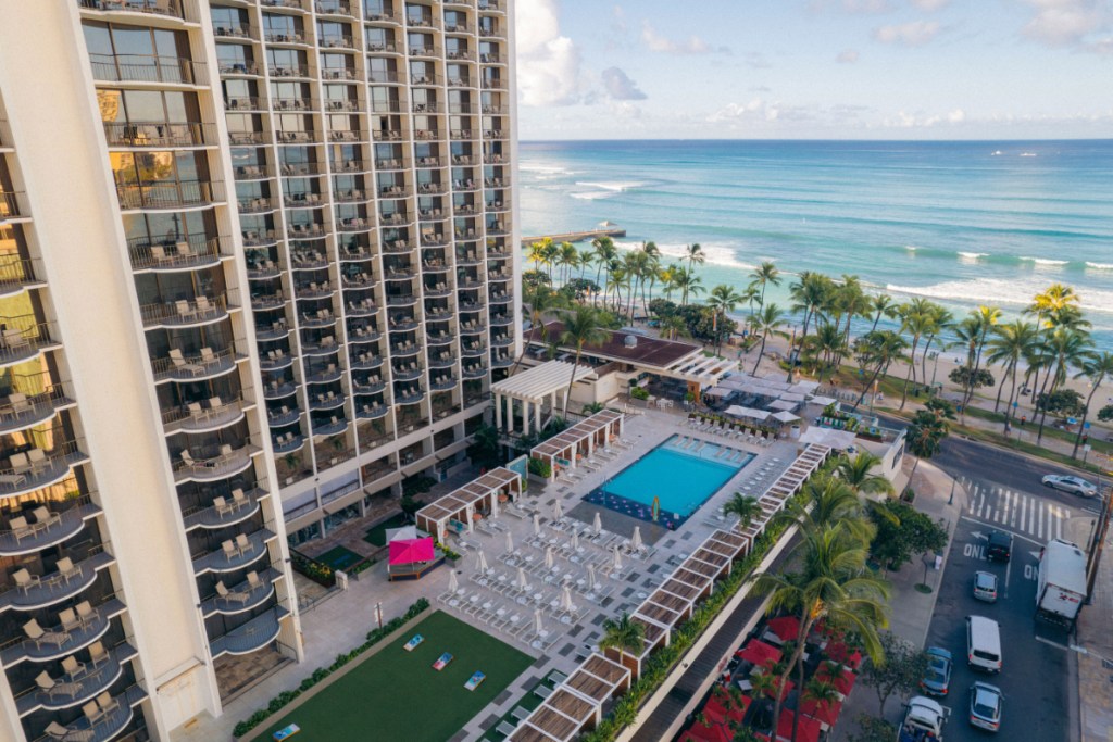 Image of a married hotel overlooking Waikiki Beach, seen from an aerial view of the hotel's pool, then the beautiful palm trees and ocean